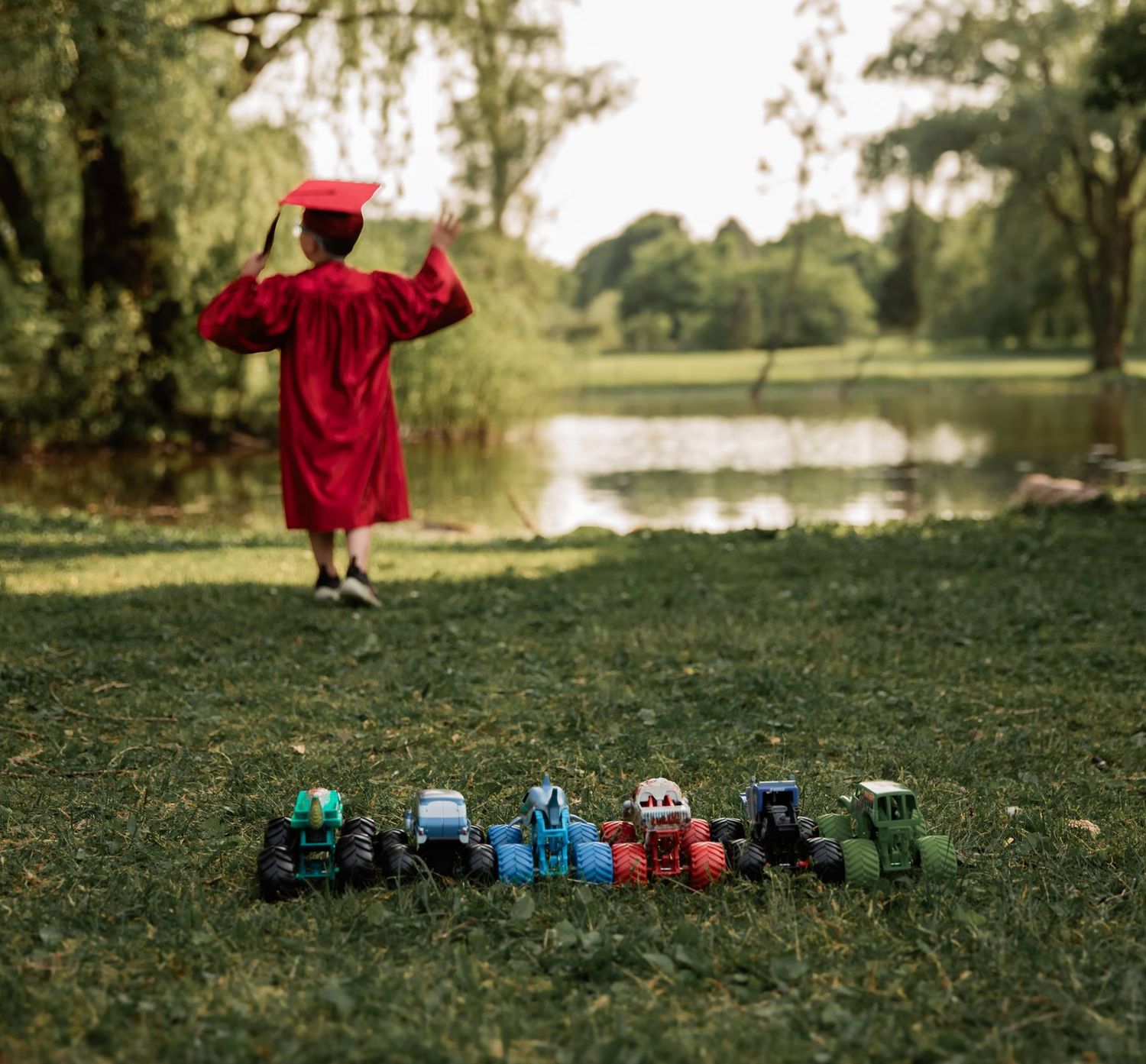 A red-robed graduate celebrates with arms raised while toy tractors line up in front on lakeside grass.