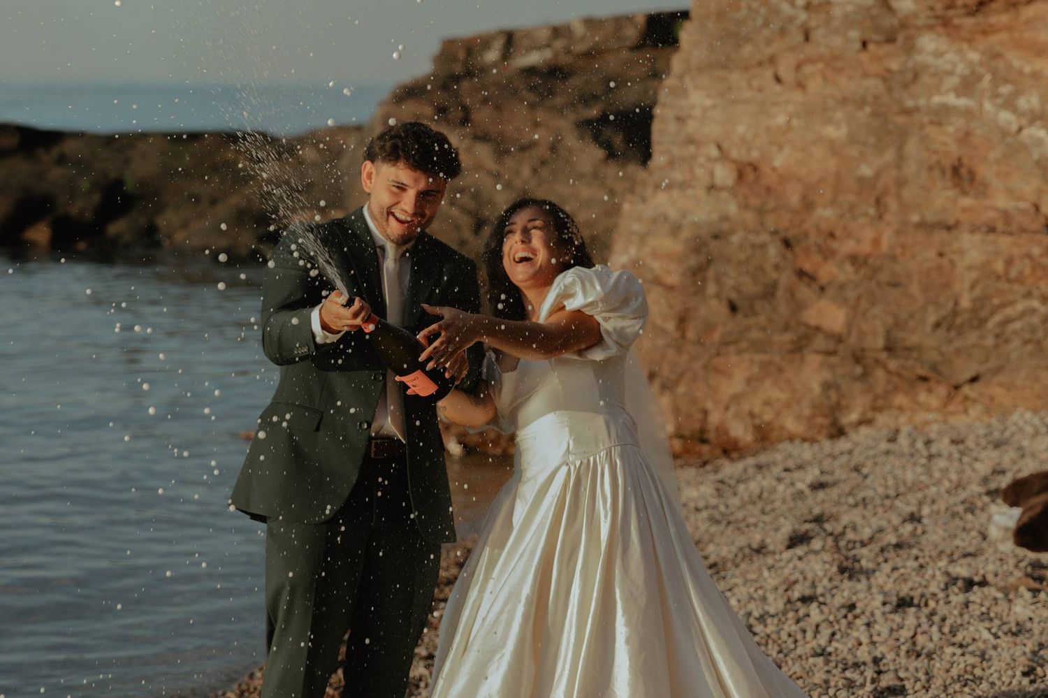 Joyful newlyweds celebrating on a rocky beach, popping champagne with water splashing around them in wedding attire.