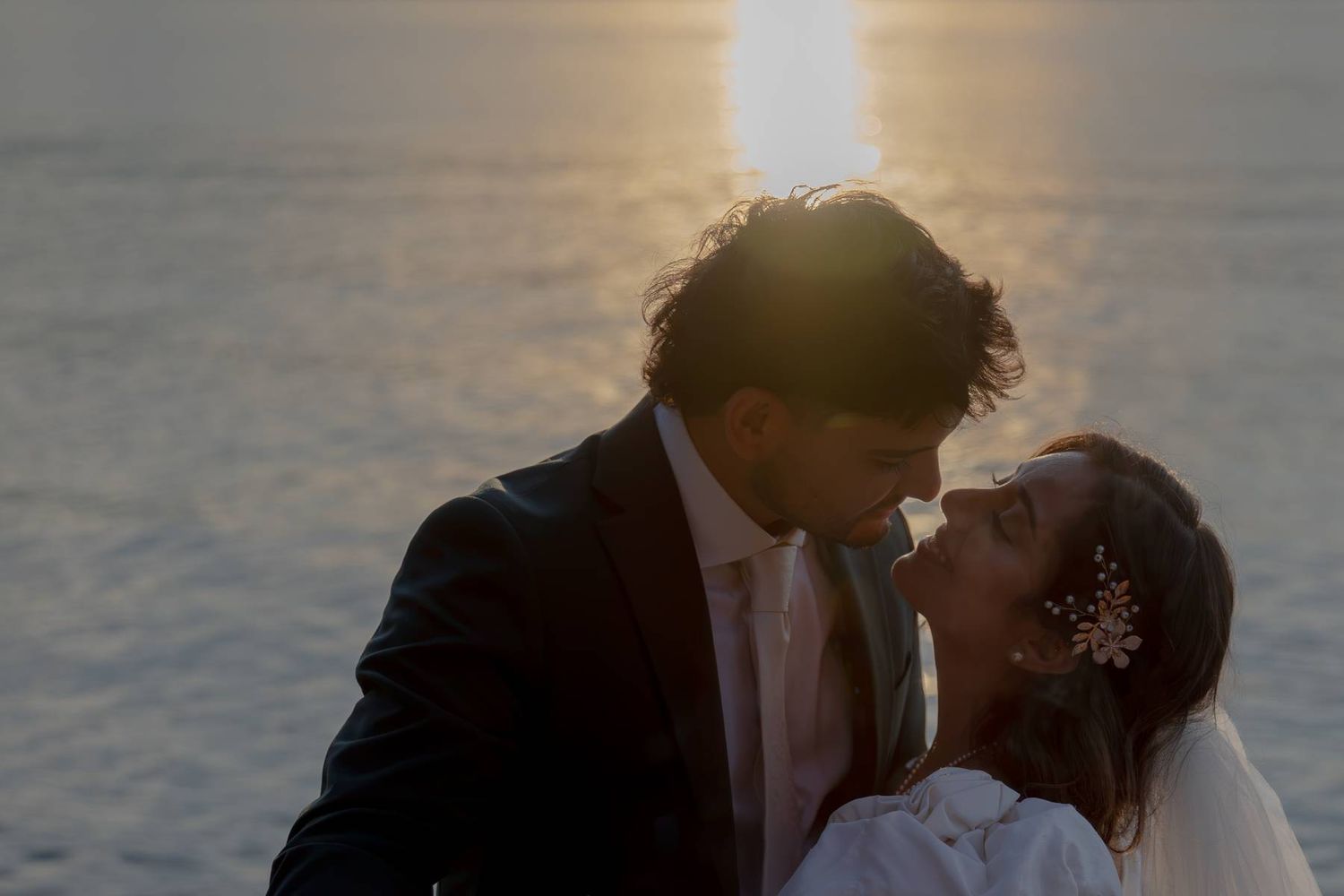 Couple sharing a romantic kiss at sunset by the ocean, bride wearing floral hair accessory and groom in dark suit.