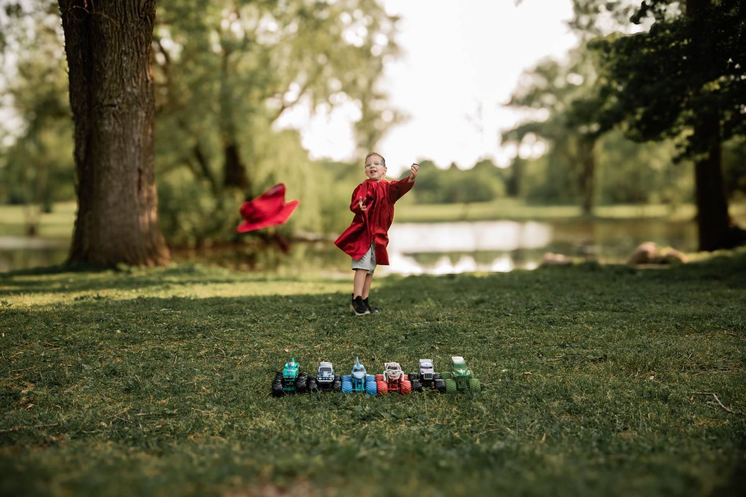 A graduate leaps joyfully in a red gown above a row of toy tractors near a serene pond.