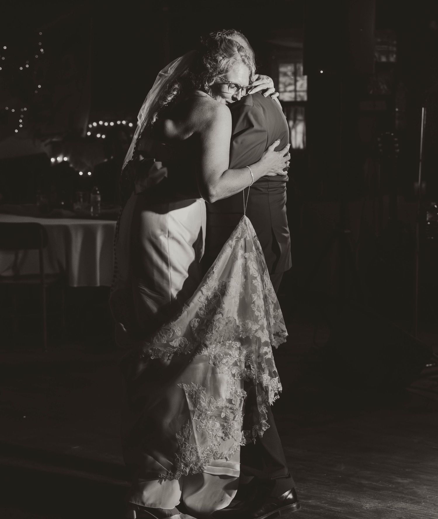 A couple shares an intimate slow dance in formal attire at a dimly lit venue with twinkling lights in the background.