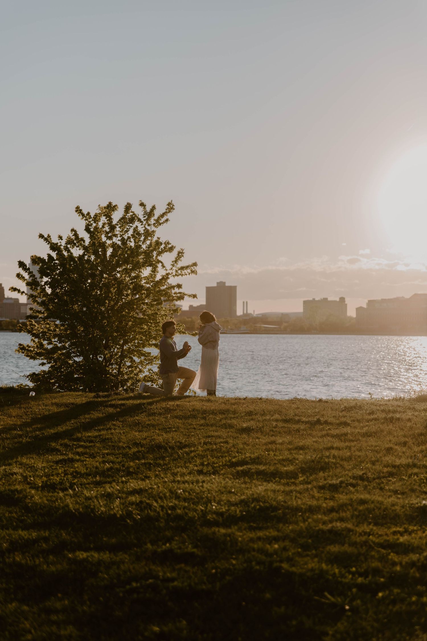 Couple sharing a romantic marriage proposal at sunset by a waterfront park with city skyline in background.
