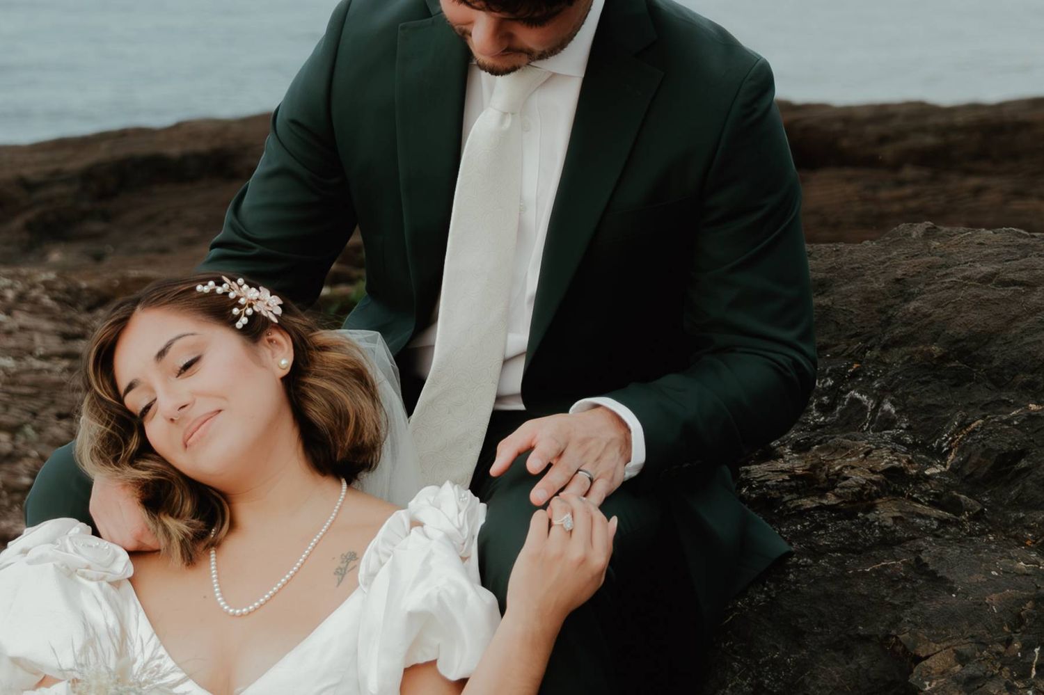 Bride in white dress and groom in green suit share a tender moment on rocky coastal cliffs.