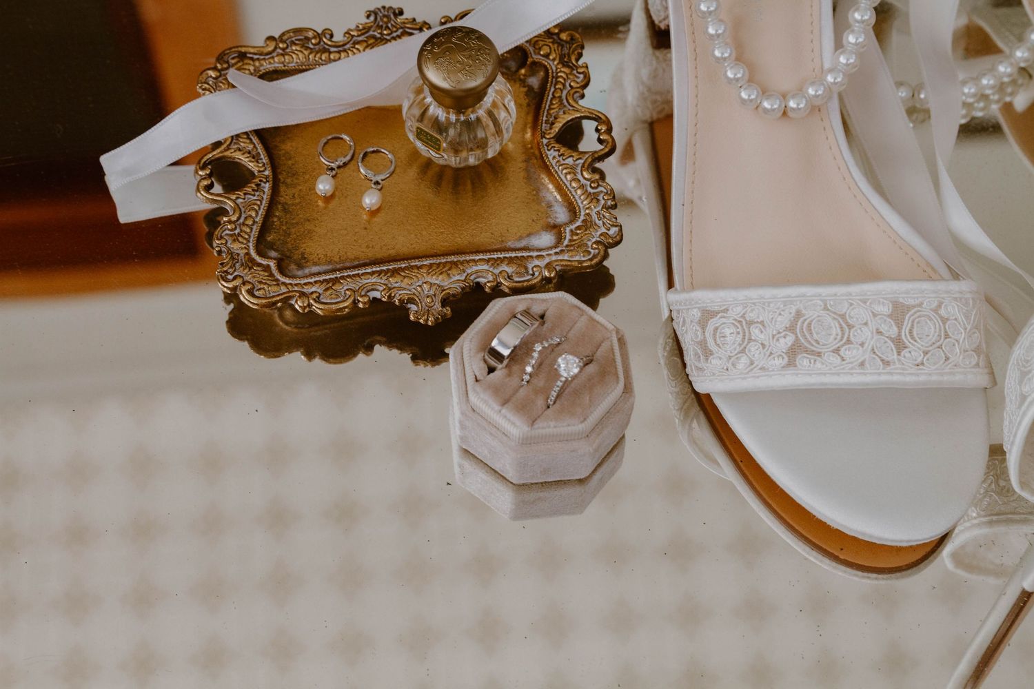 Elegant wedding accessories including a vintage mirror tray, pearl necklace, and delicate perfume bottle on white lace.