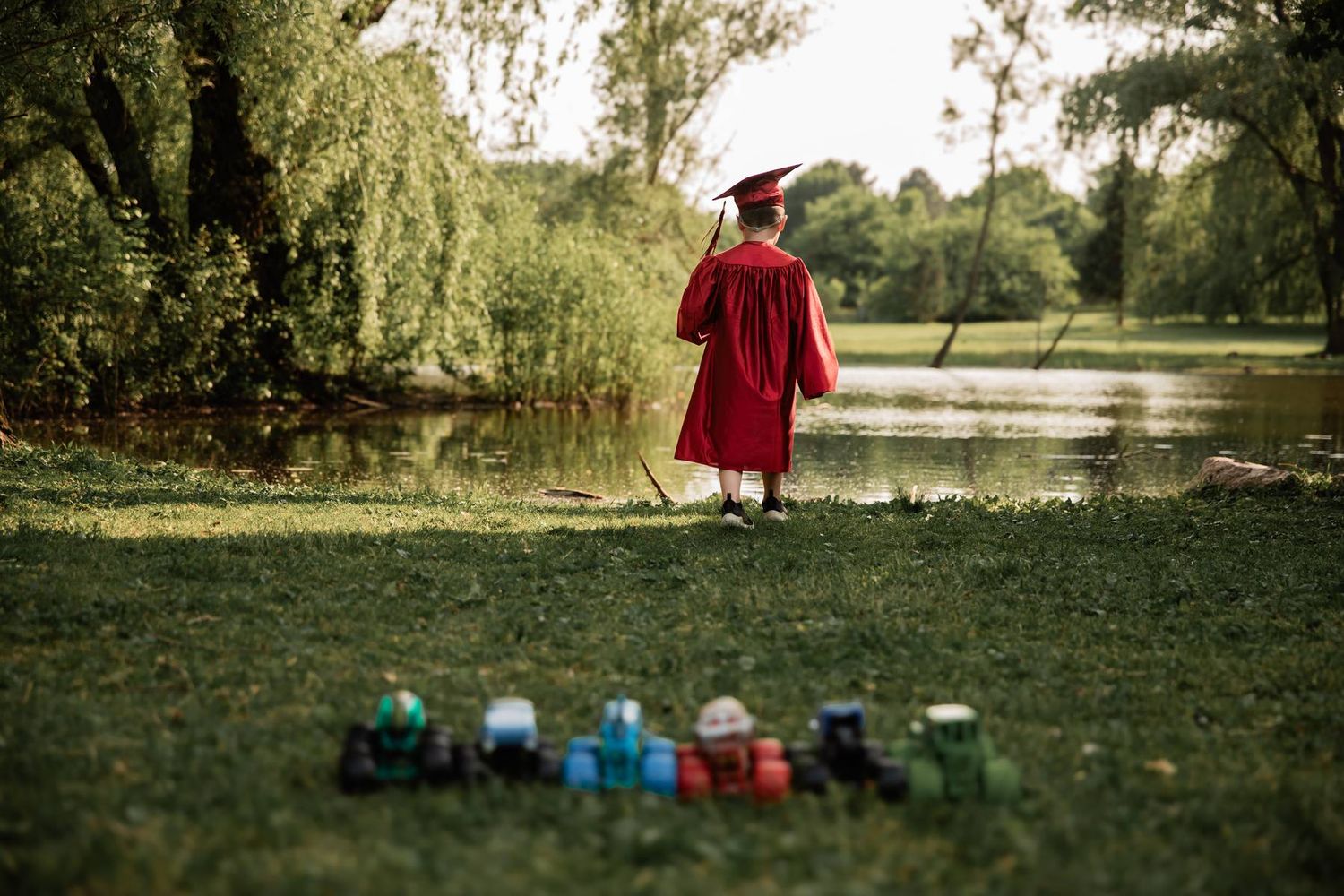 A small graduate in red cap and gown stands by a lake with colorful toys lined up on grass.