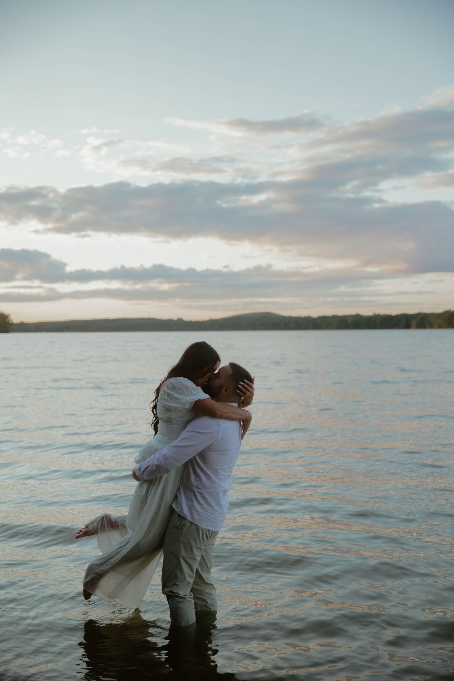 A couple embraces while standing in shallow water at sunset with a serene lake and cloudy sky in the background.