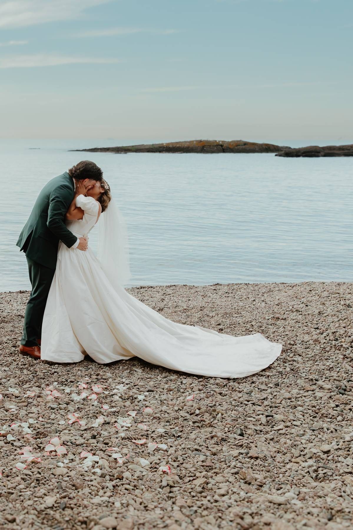 Bride and groom share a romantic kiss on a rocky beach shoreline, with calm blue ocean waters in the background.