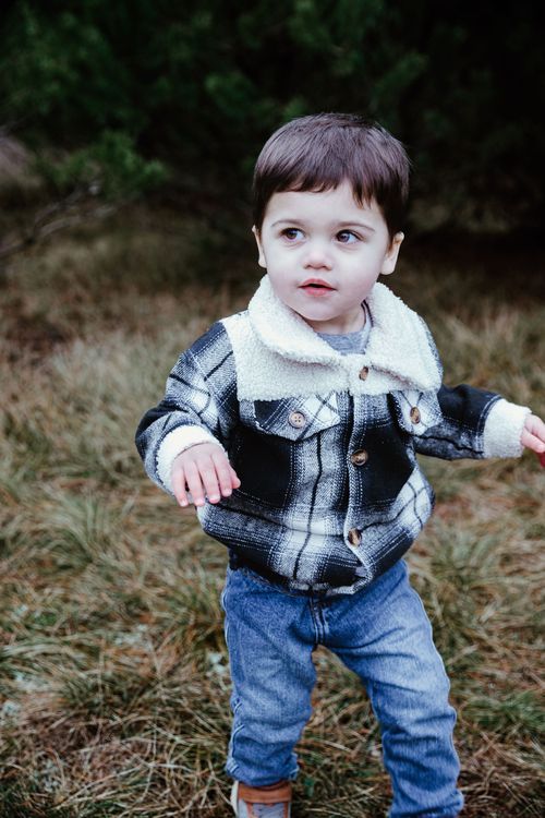 A toddler wearing a plaid jacket and jeans stands in front of autumn foliage.