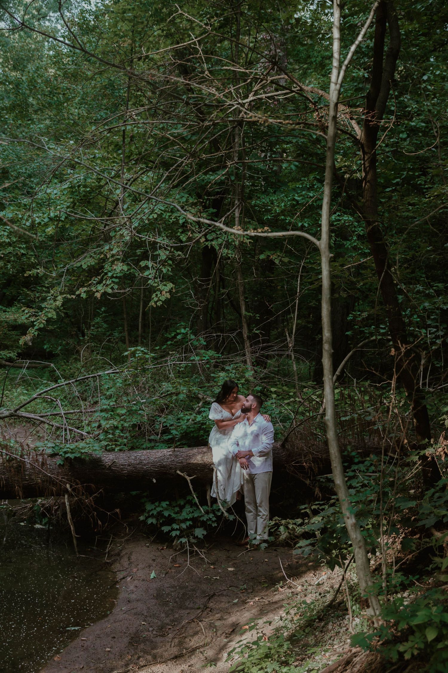A couple embraces on a wooded trail surrounded by lush green forest and natural scenery.
