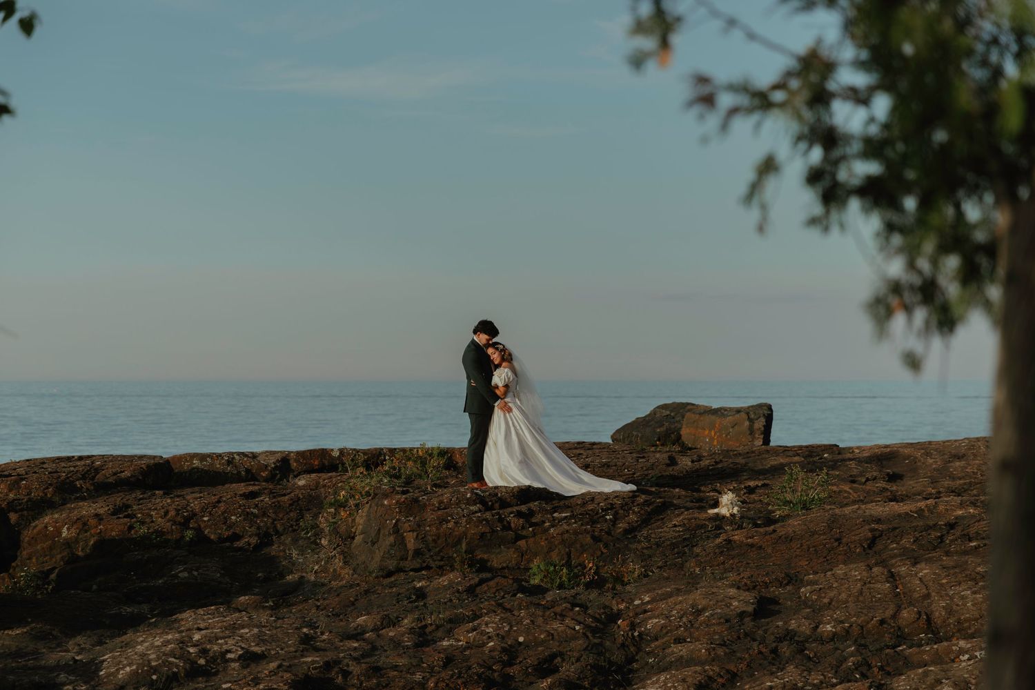 Bride and groom embrace on rocky cliffside overlooking calm ocean at sunset in elegant wedding attire.