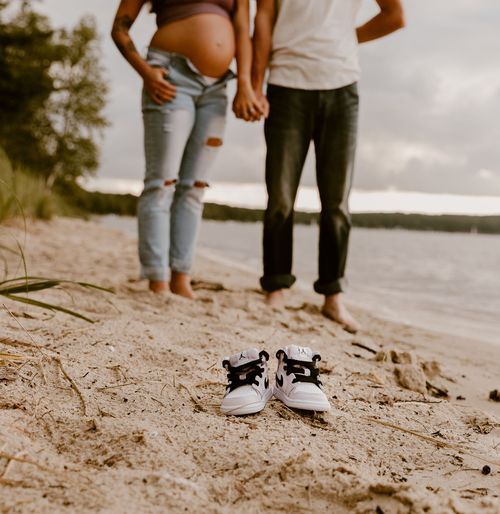 Tiny baby shoes placed on sandy beach with couple's feet in background.