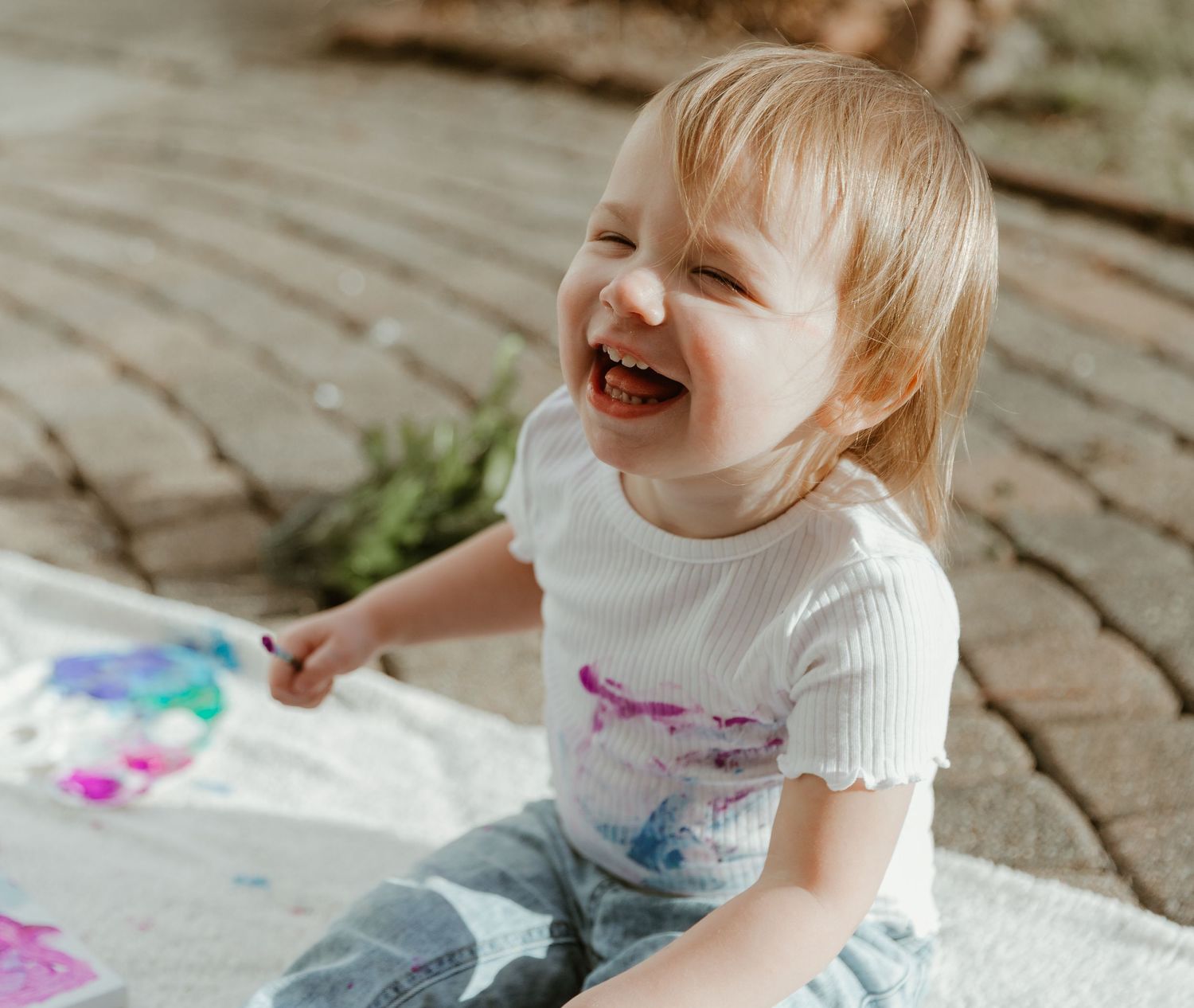 A cheerful toddler in a white shirt laughs while sitting on brick pavers during an outdoor art activity.