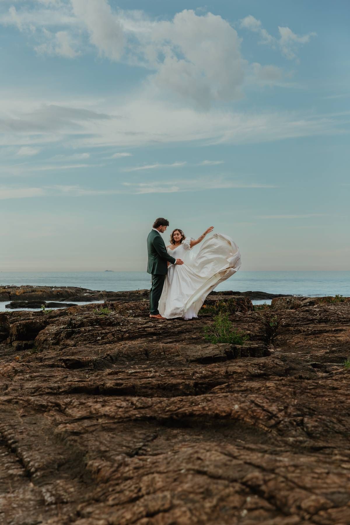 Bride and groom stand on rocky coastal cliffs, her white gown flowing in the wind beneath a blue sky.