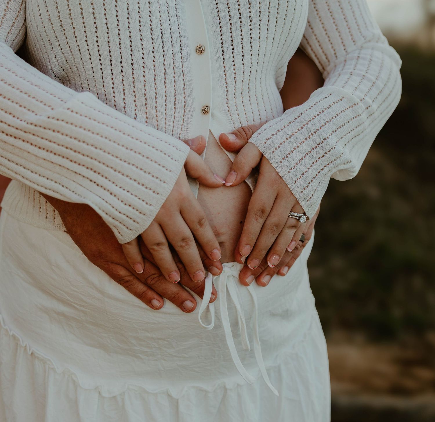 Close-up of hands with engagement ring against a white knit sweater and flowing skirt in soft natural light.
