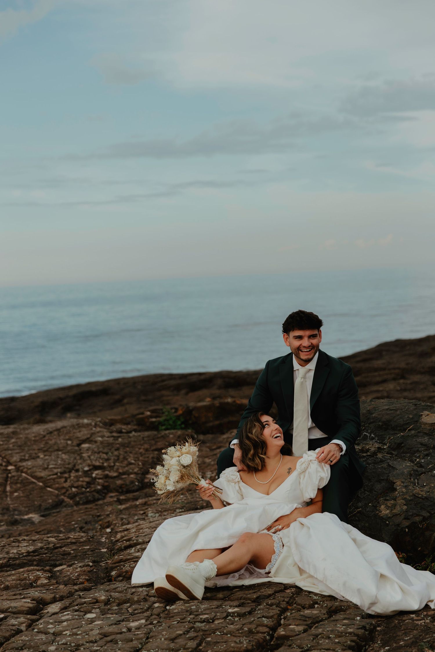 Bride and groom sitting on coastal rocks, laughing joyfully, bride in white gown holding bouquet, groom in dark suit, ocean backdrop.