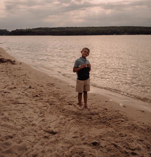 A person stands on a sandy beach at sunset with a serene lake and wooded shoreline in the background.