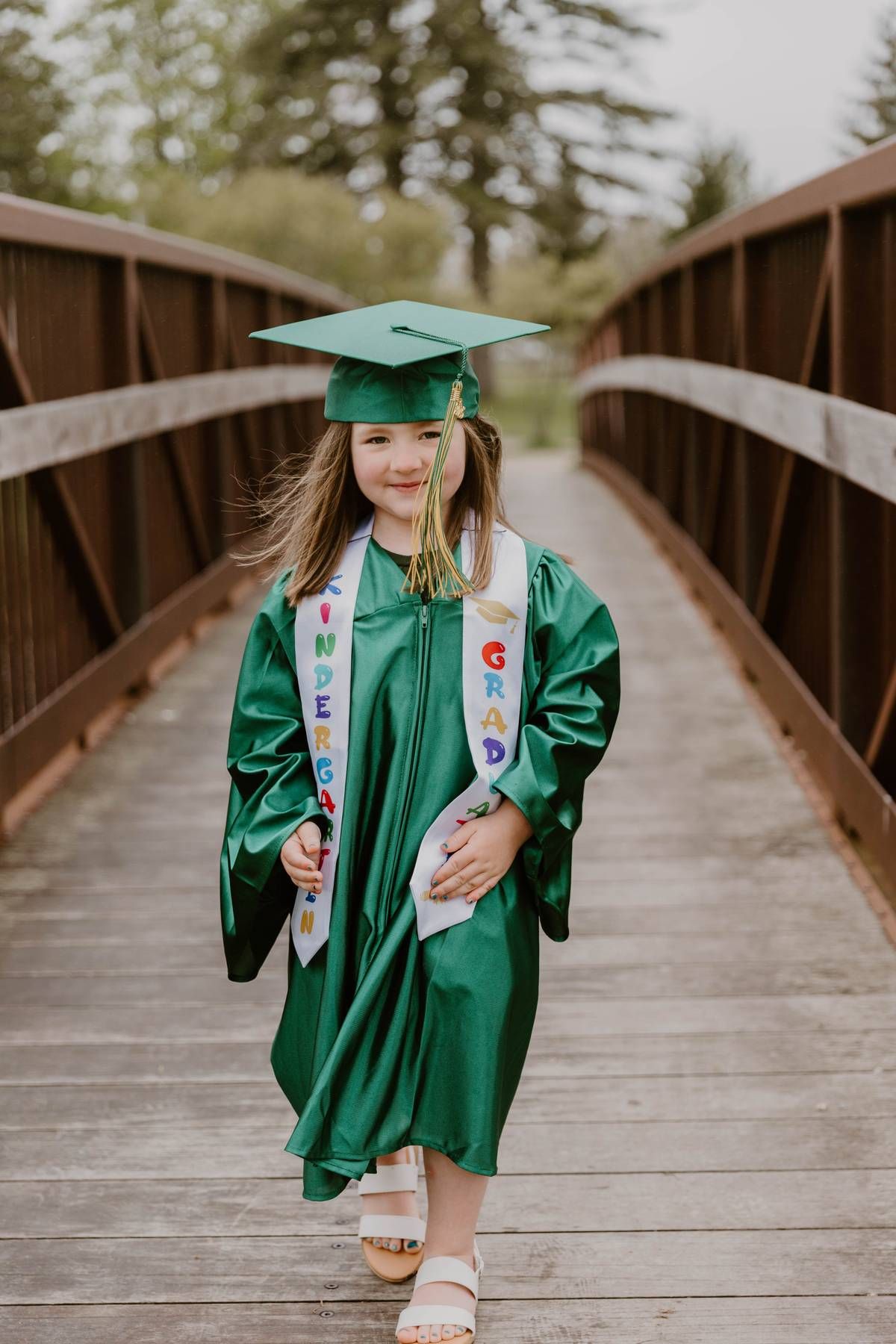 Graduate in emerald green cap and gown walks across wooden bridge in outdoor setting.
