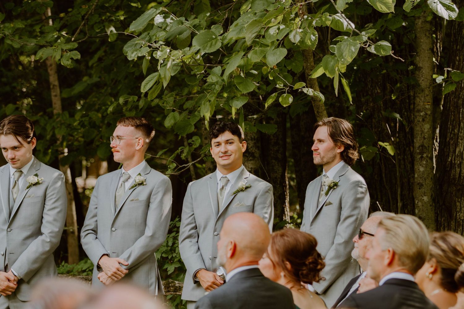 Wedding party members in gray attire gather outdoors under green foliage for formal photos.