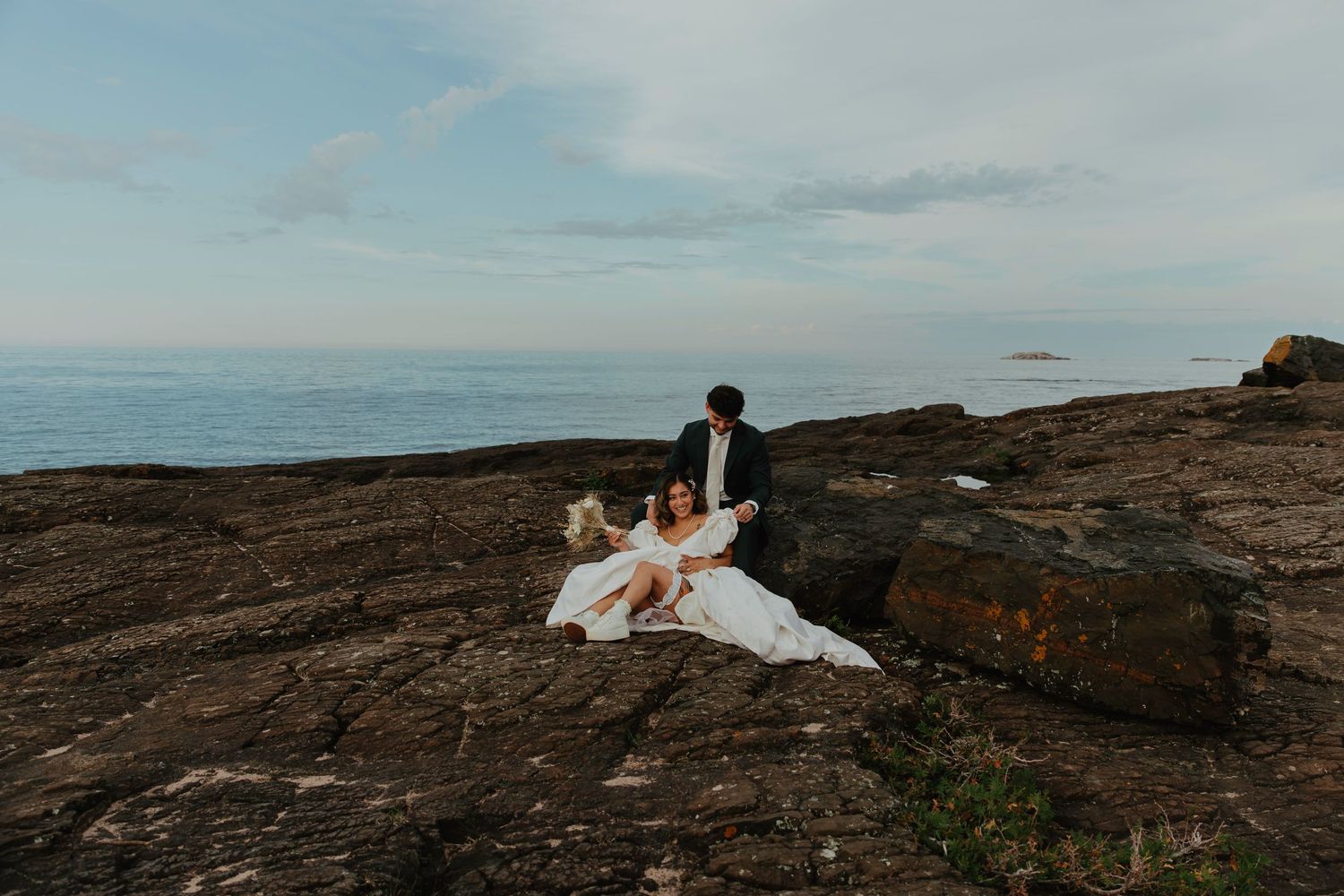 Bride and groom sitting on rocky coastal cliffs with ocean horizon backdrop during their outdoor elopement ceremony.