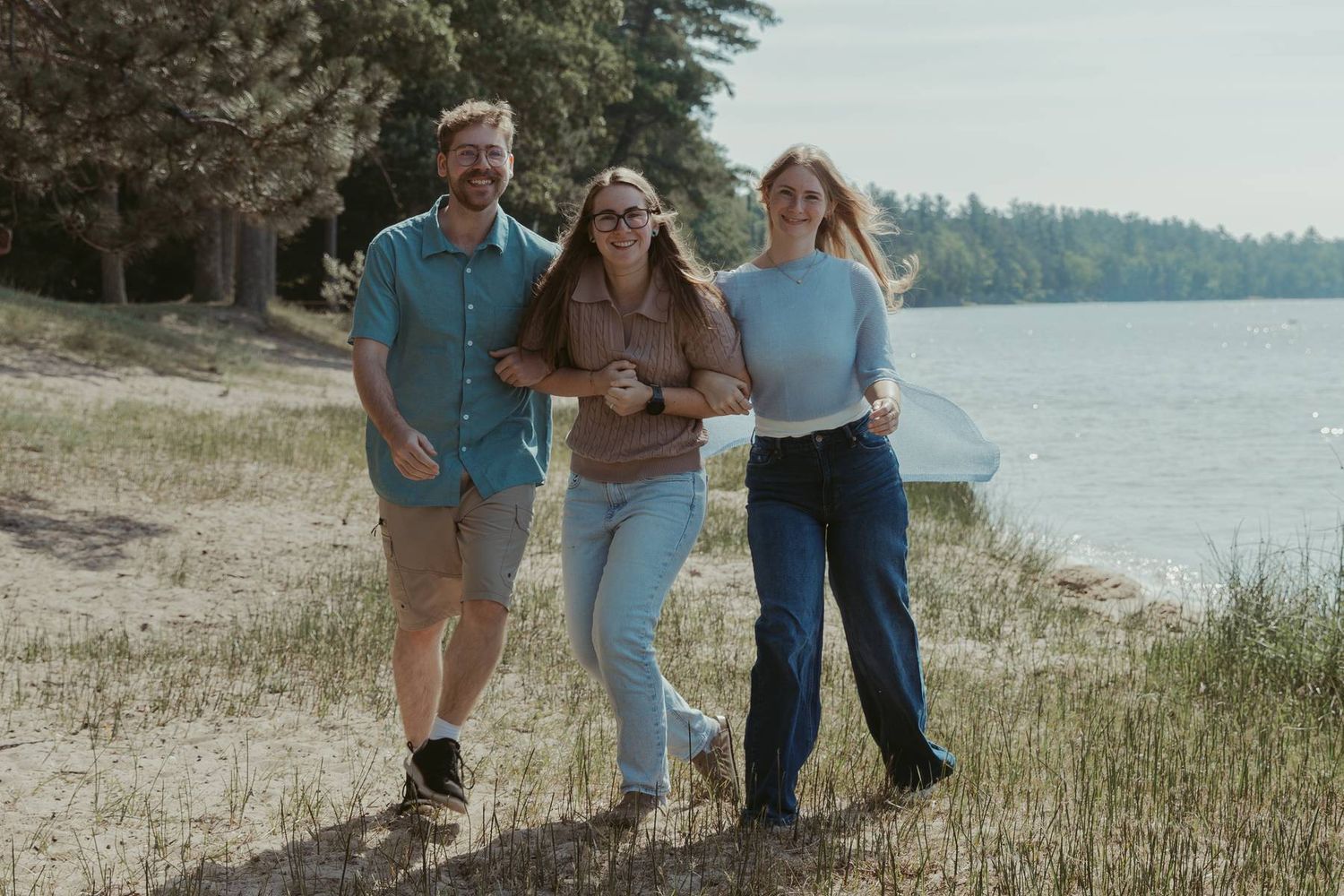 Three friends smiling and walking together along a scenic lakeside trail surrounded by green trees.