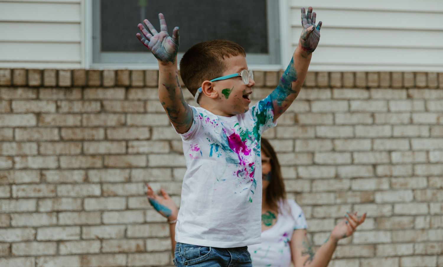 A person joyfully jumping with colorful paint-stained clothes against a white brick wall background.