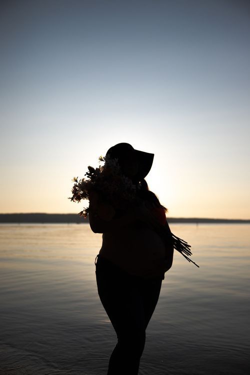 Silhouette of pregnant woman holding flowers against serene waterfront sunset.