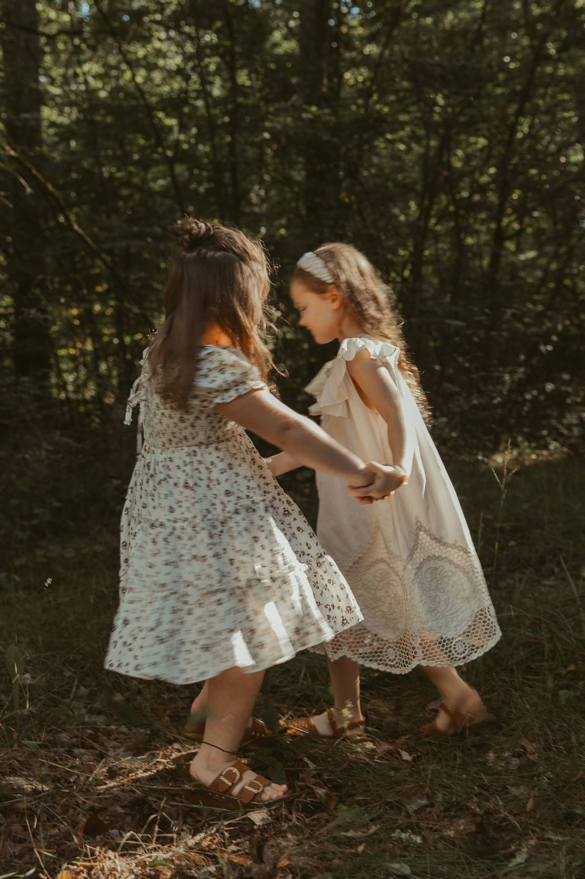 Two young girls in floral and white dresses hold hands and dance together in a sunlit forest.
