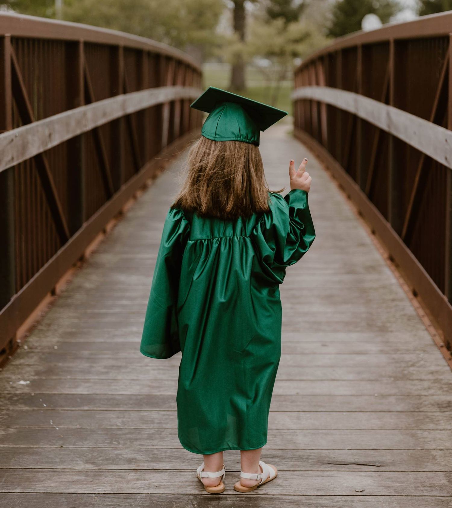 Graduate in emerald green cap and gown walking across wooden bridge during outdoor photoshoot.