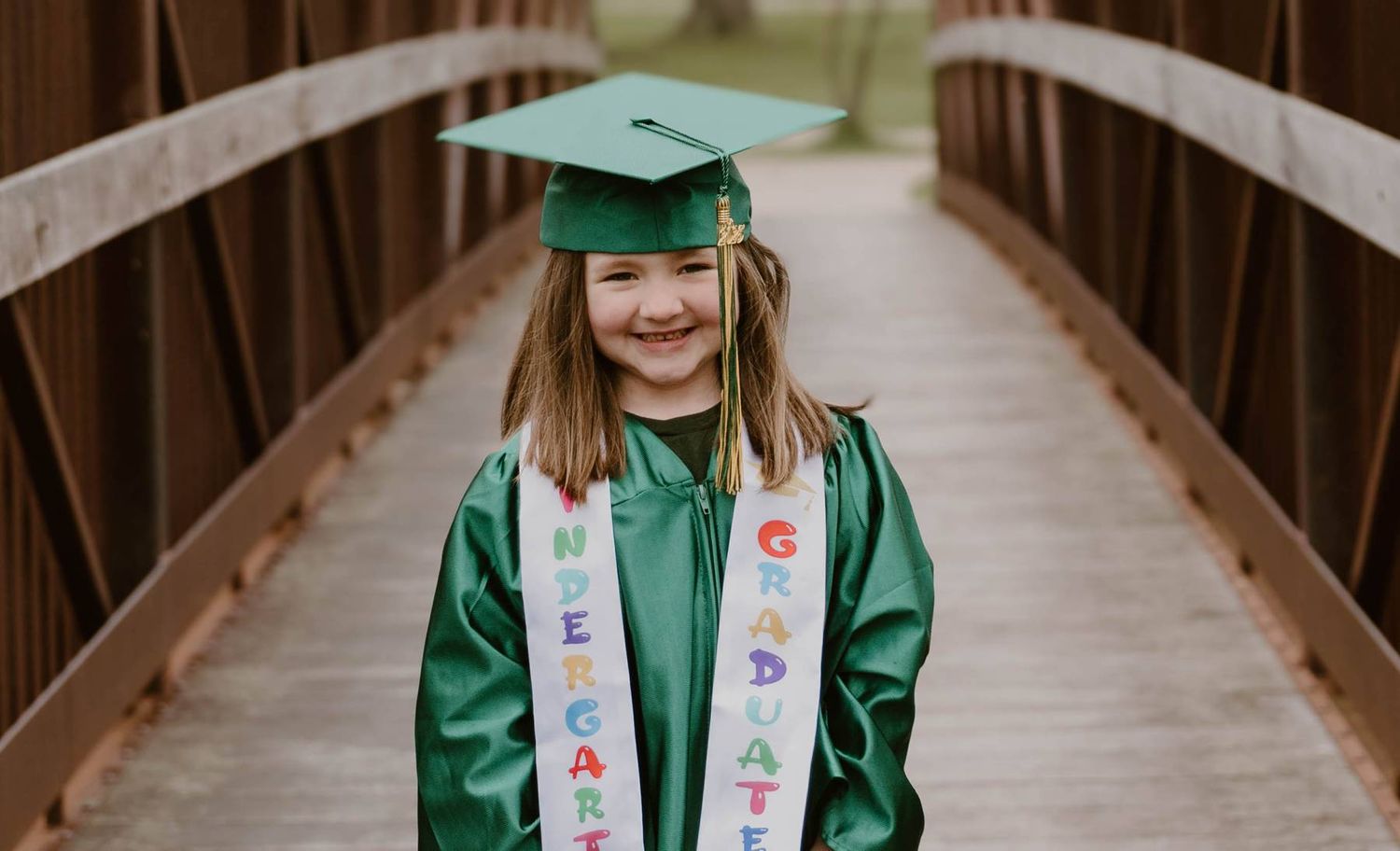 Graduate in green cap and gown wearing decorated sash stands smiling on wooden bridge.