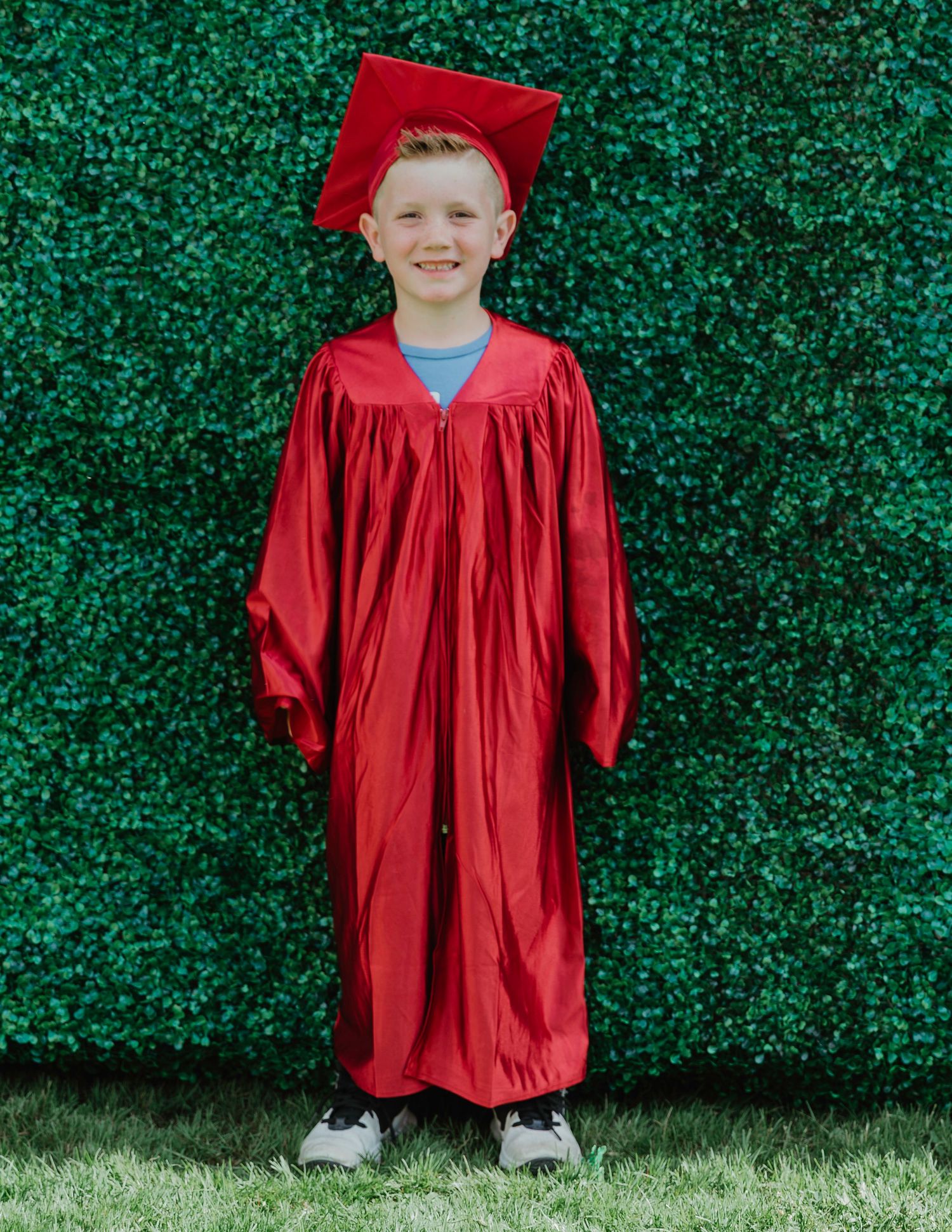 Young boy smiling in red graduation cap and gown standing in front of green hedge backdrop.
