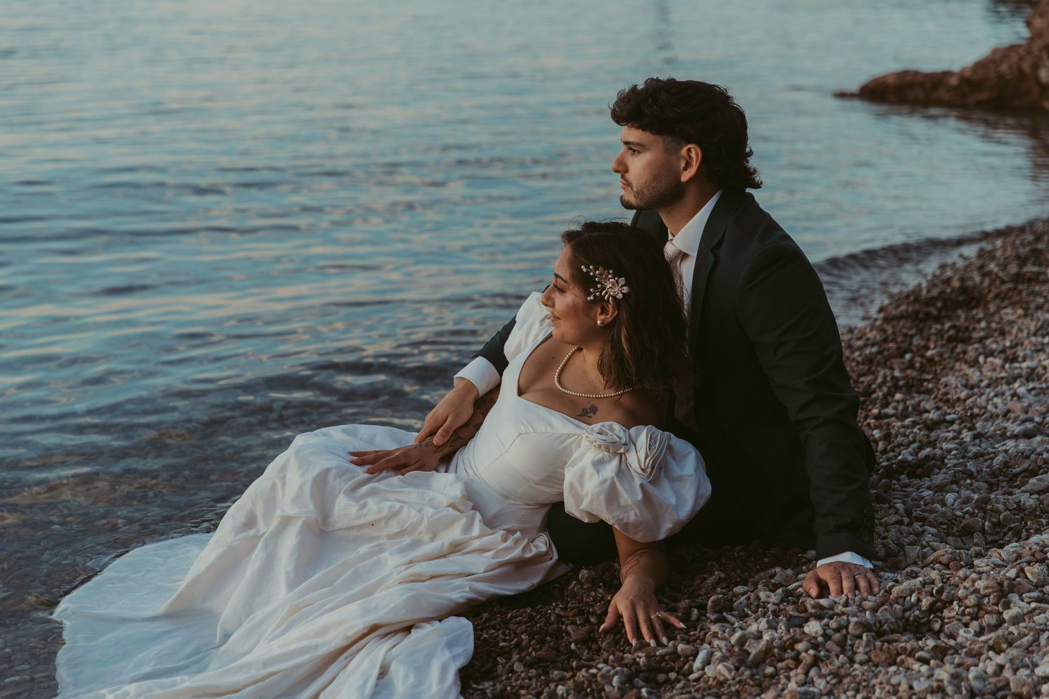 A bride and groom sit together on a rocky shoreline at sunset, the bride in a white gown with puff sleeves and floral crown.