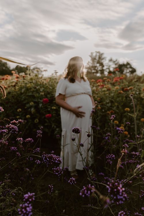 A pregnant figure in a white dress stands among colorful wildflowers at golden hour.