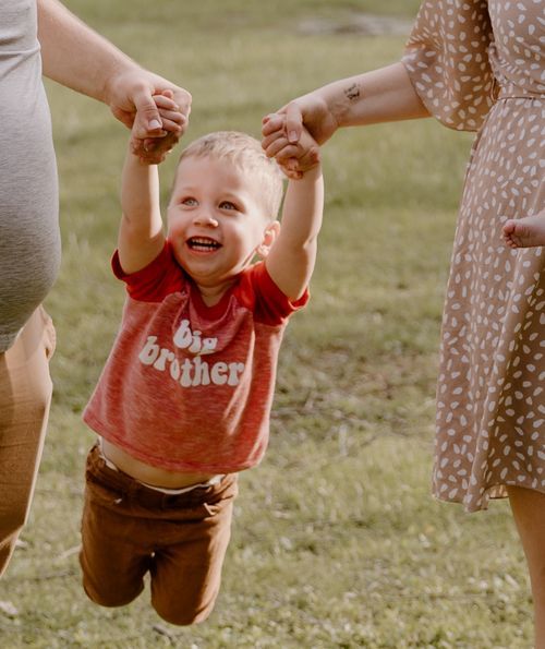 Young child in red shirt being lifted by hands while laughing outdoors.