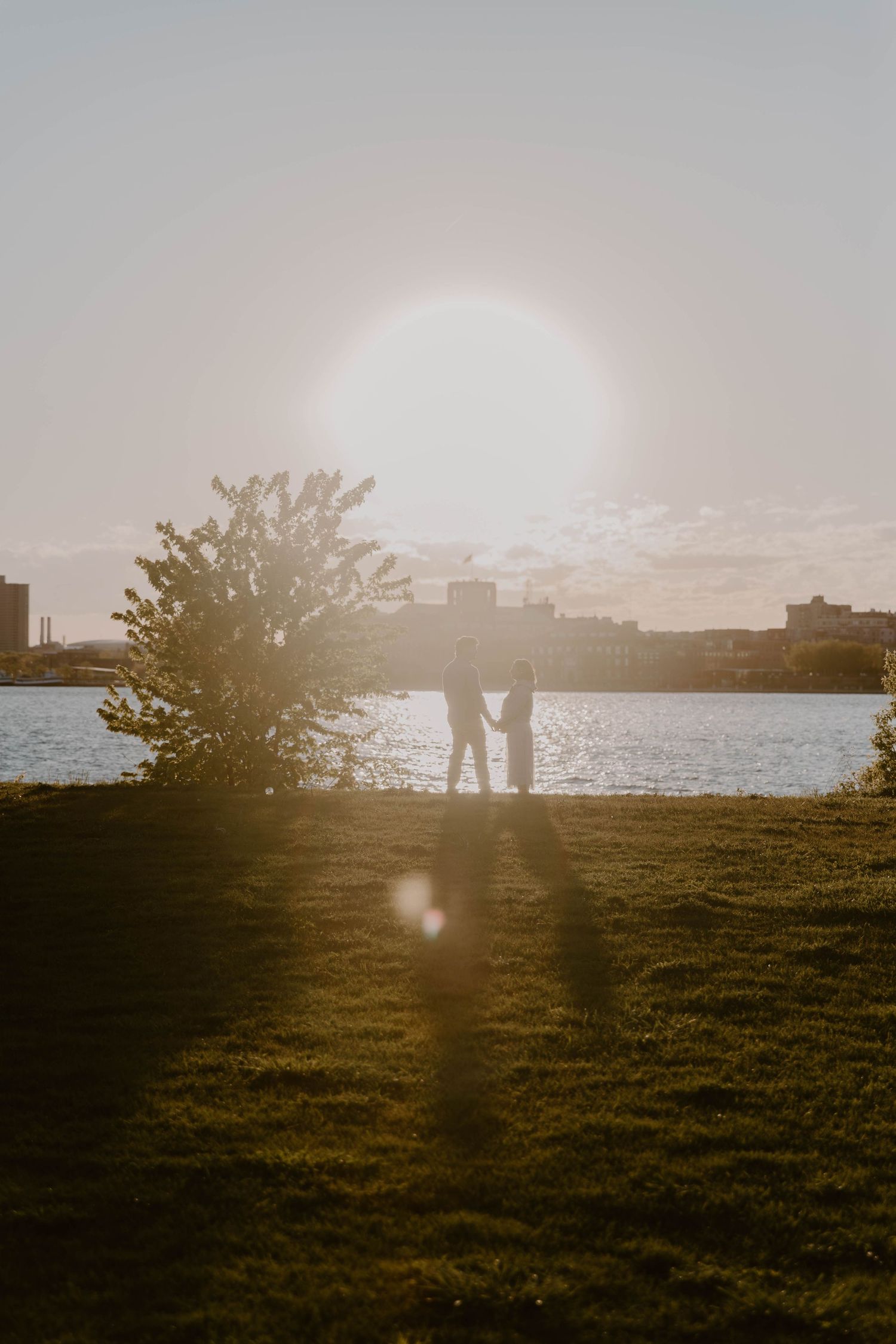 Silhouette of couple holding hands at golden sunset by a river, with lens flare and lush green grass.