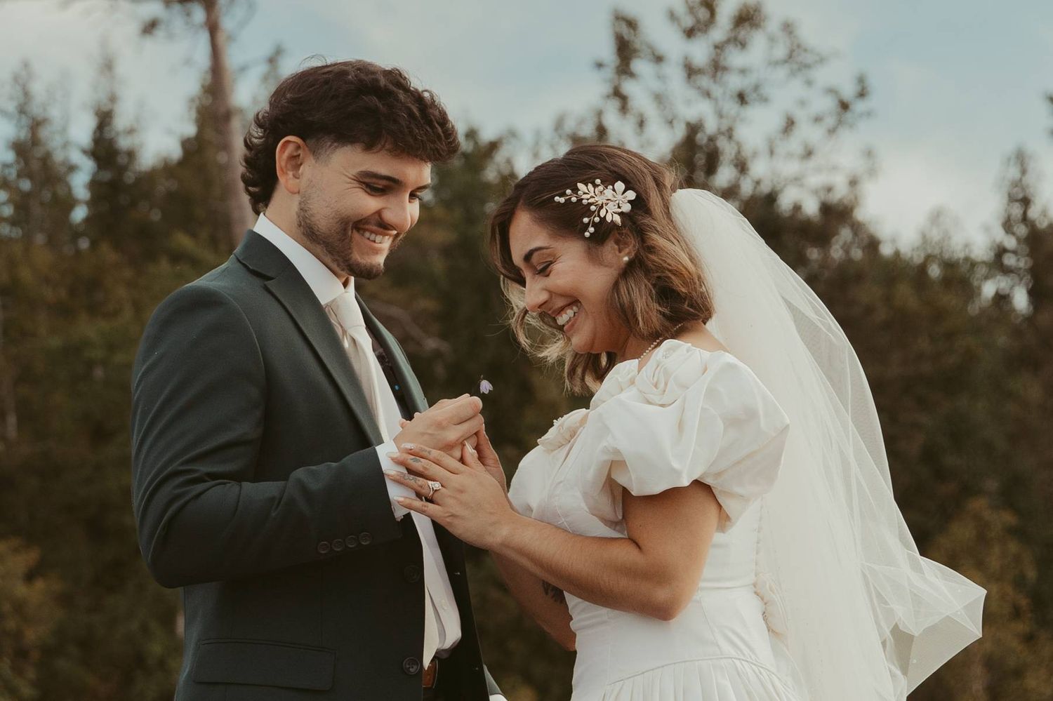 A smiling bride and groom hold hands outdoors on their wedding day, with trees in the background.