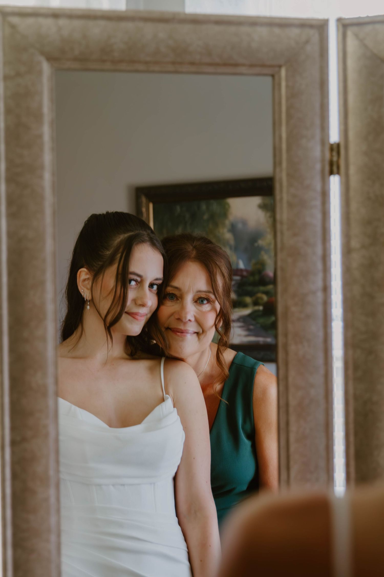 A tender moment reflected in a mirror shows two figures in white and green attire sharing an intimate embrace.