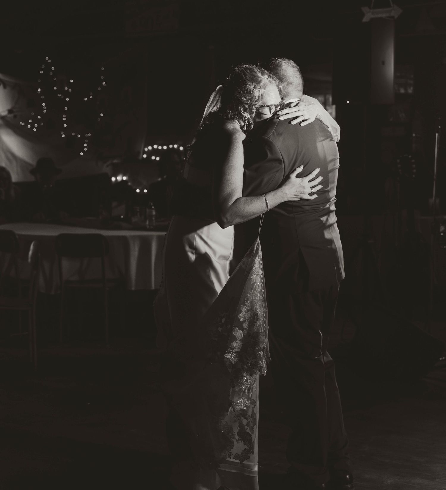 Two people slow dancing together in a dimly lit setting with string lights visible in the background.