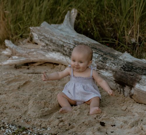 Baby in gray dress playing near weathered driftwood at beach.