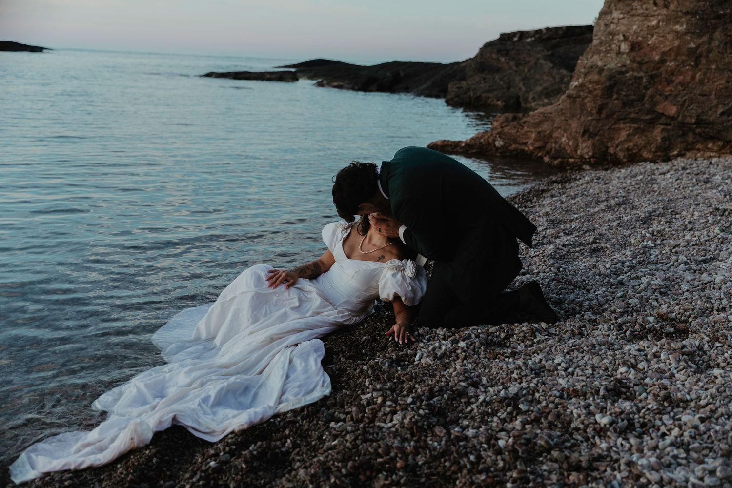Bride in white gown and groom in dark suit share romantic kiss on rocky beach at dusk near calm ocean waters.