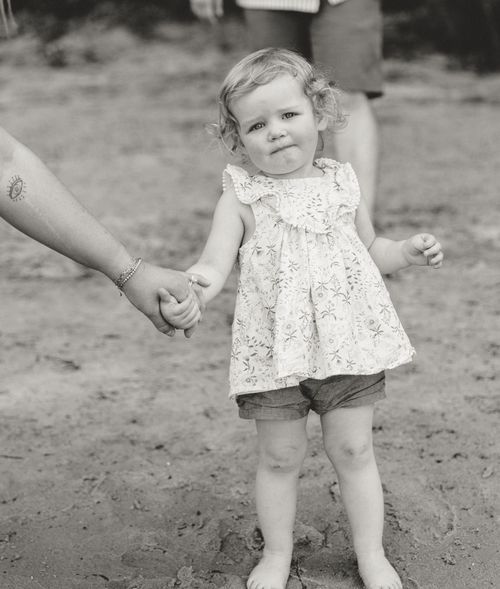 A small child in a floral dress holding an adult's hand while standing on dirt ground.
