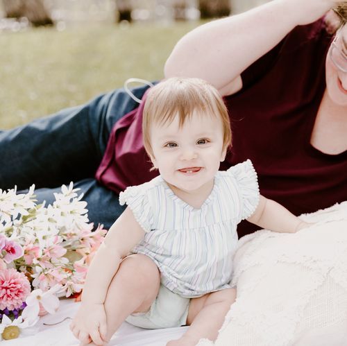 Baby in white ruffled outfit sitting on blanket with pink flowers.