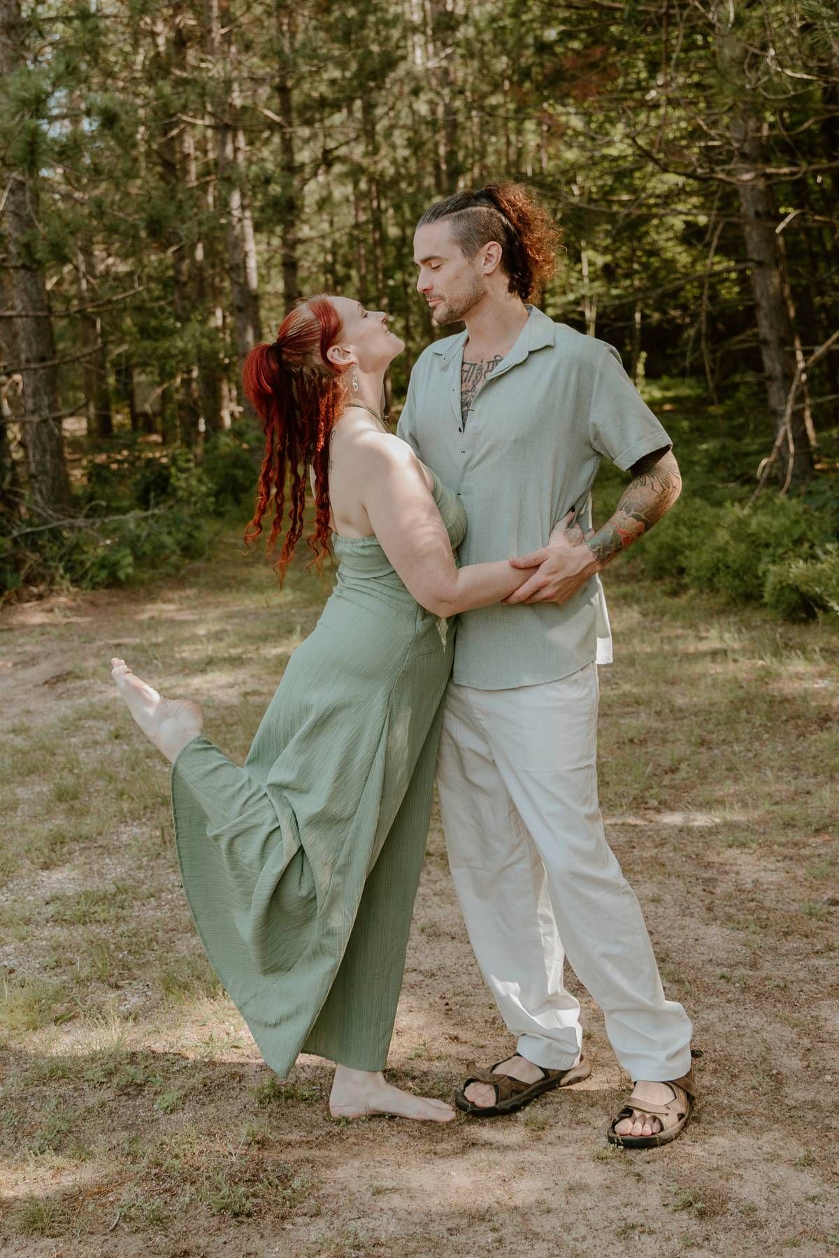 A couple in matching sage green outfits dancing romantically in a forest clearing, woman with red dreadlocks.