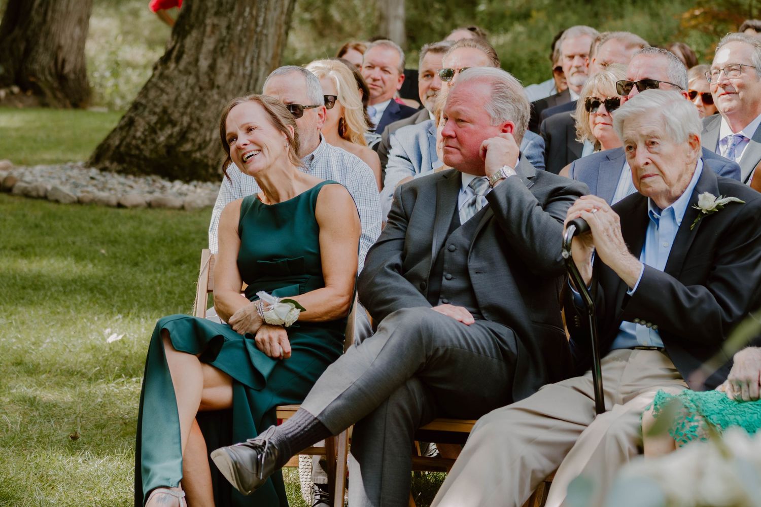 Guests seated in wooden chairs share emotional reactions during an outdoor wedding celebration.