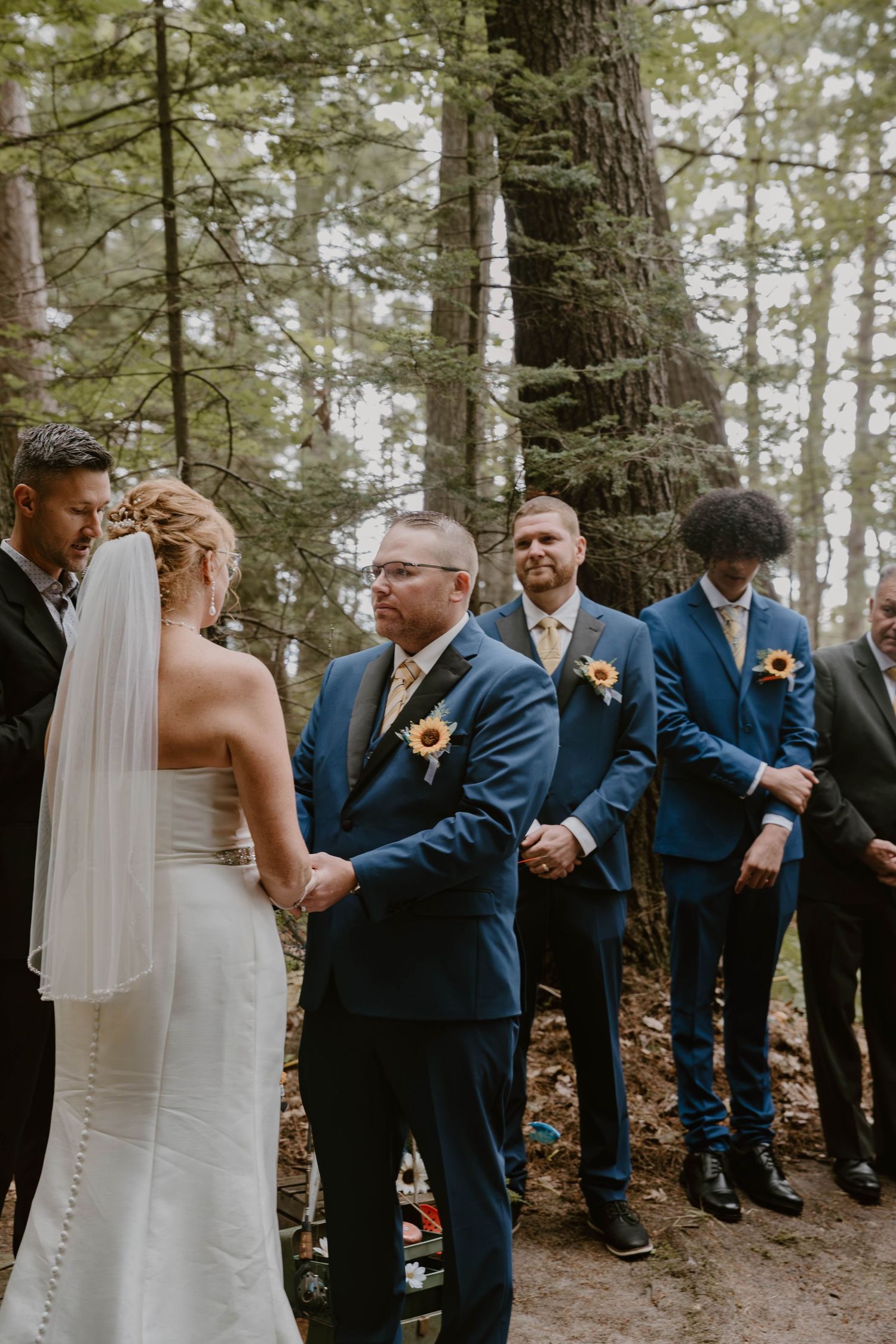 Wedding party in navy blue suits gathers in a forest setting during an intimate outdoor ceremony.