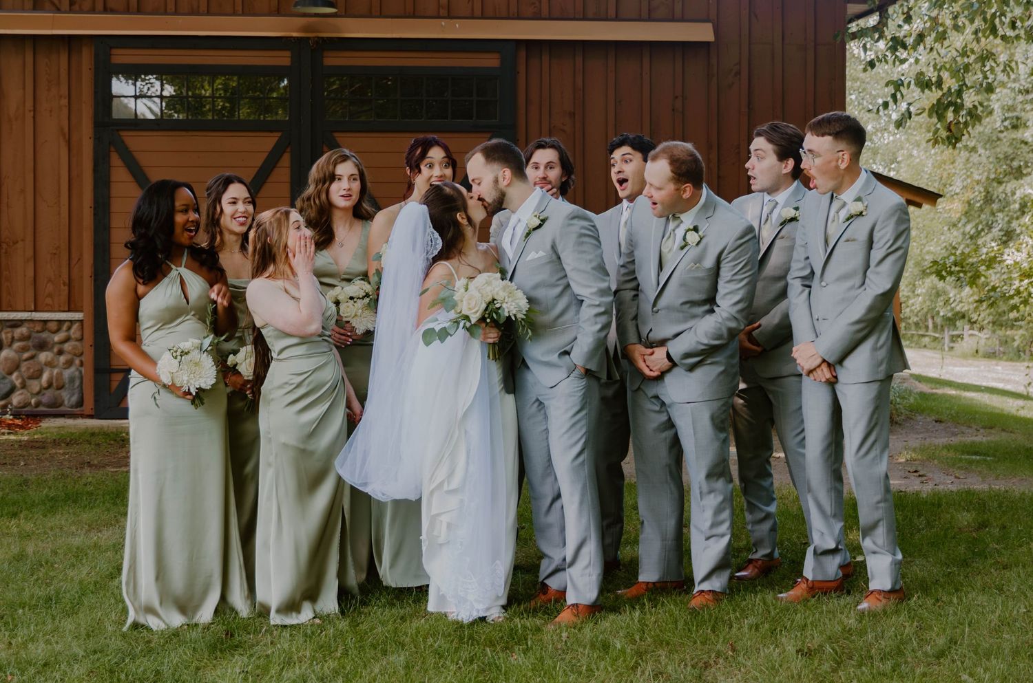 Wedding party in sage green and gray attire poses together on grass in front of rustic wooden barn wall.