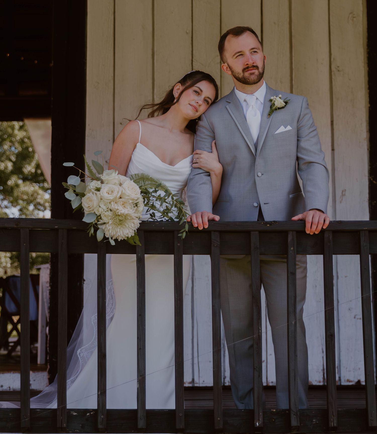 A couple stands on a rustic wooden porch with white floral bouquet during their wedding day.