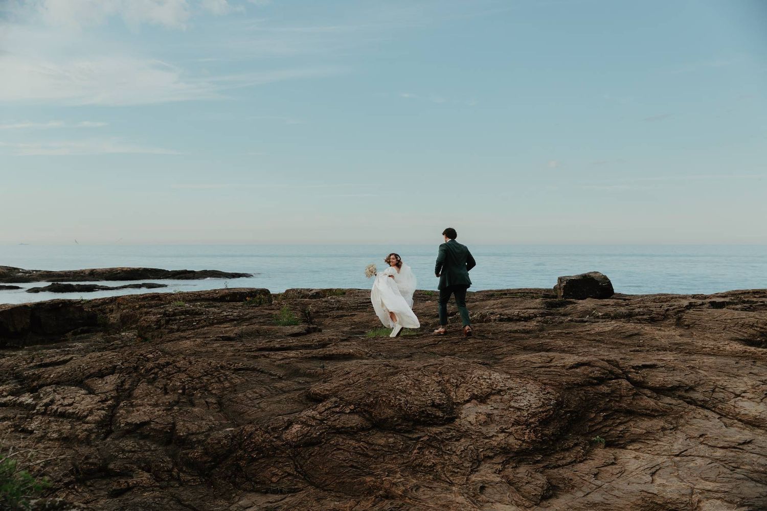 A bride in a white gown and groom walk along rocky coastal cliffs with the ocean horizon under a clear blue sky.