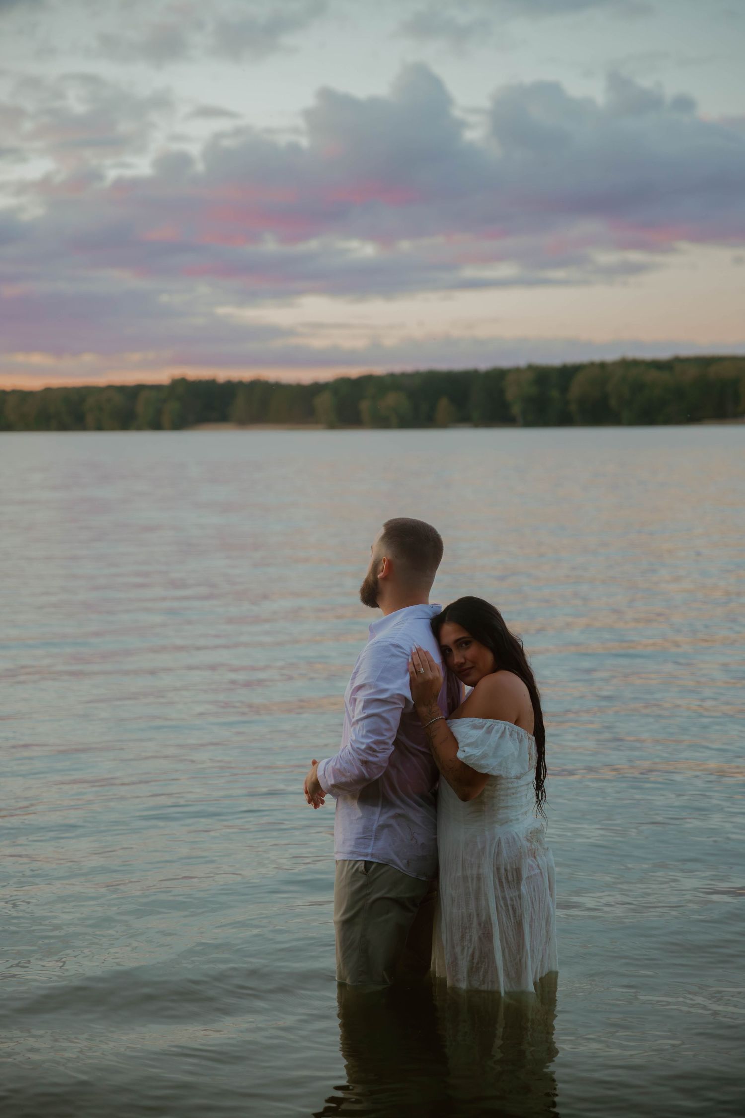 A couple stands together in a lake during sunset with dramatic pink and purple clouds in the sky.