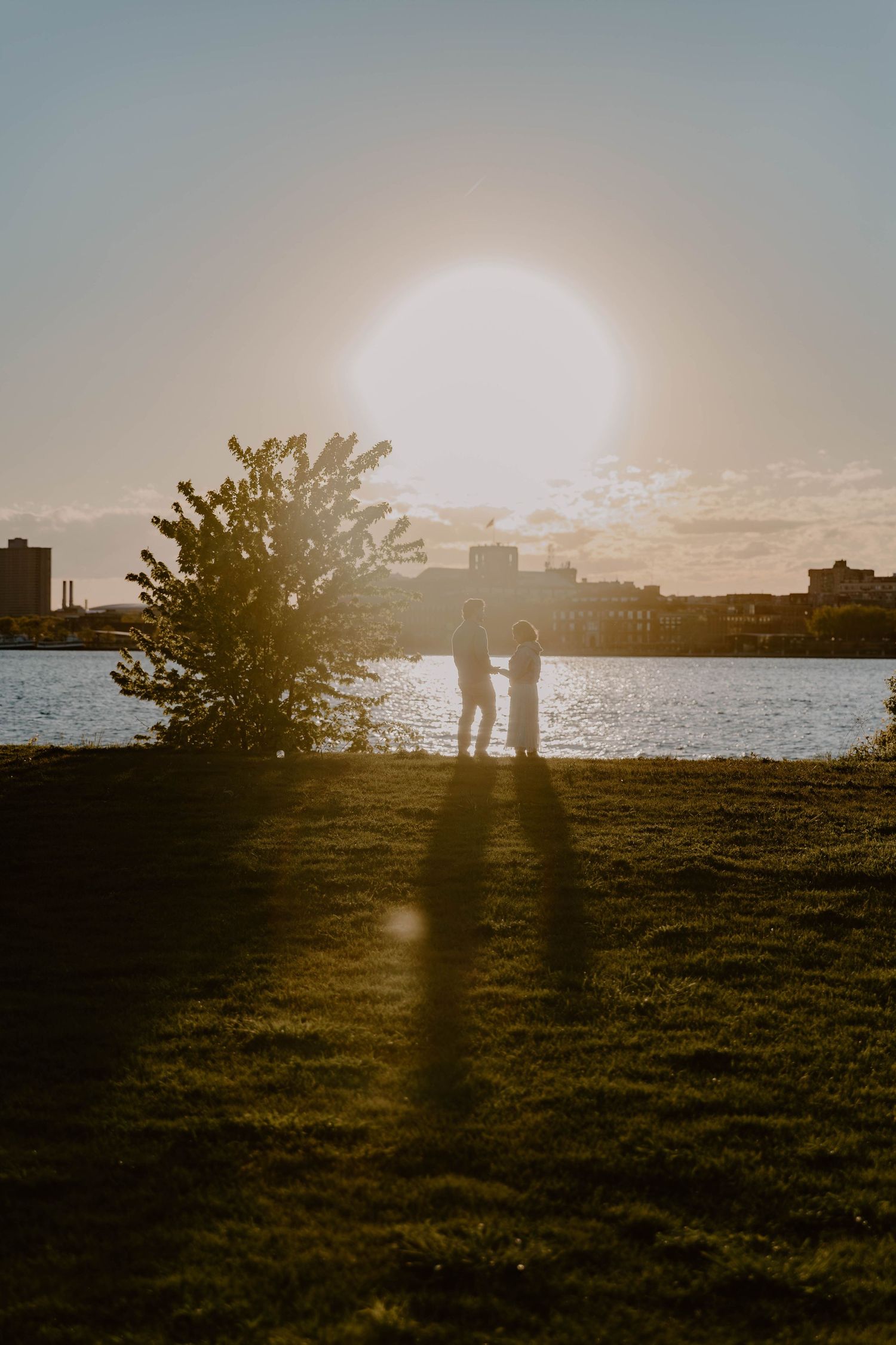 Couple silhouetted against golden sunset by waterfront park with city skyline in background.