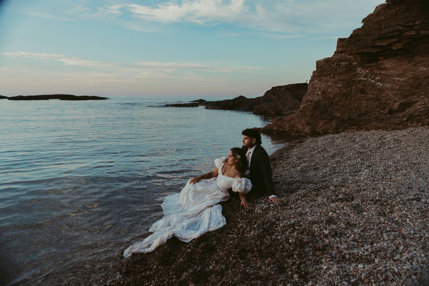 Bride in white gown and groom sit romantically on rocky shoreline at sunset with calm blue sea and cliffs behind them.